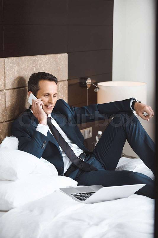 Smiling businessman in formal suit sitting on hotel room bed while talking on phone, stock photo