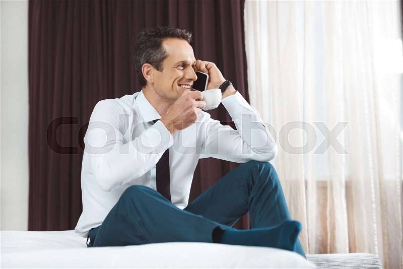 Businessman in formal wear sitting on bed in hotel room, talking on phone and drinking coffee, stock photo
