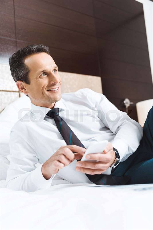 Businessman in formal wear lying on bed in hotel room and using his smartphone, stock photo