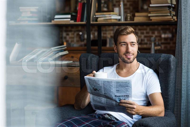 Smiling young man in home clothes sitting in armchair and reading a newspaper, stock photo