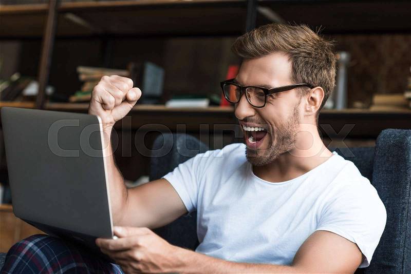 Young man sitting with laptop in armchair and cheering with his fist as he is looking at computer screen, stock photo