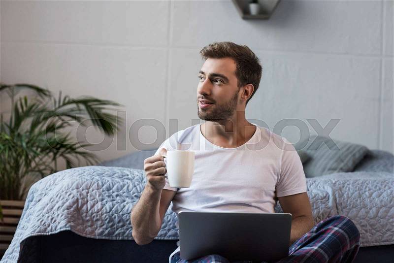 Young man sitting on floor in his bedroom with laptop and cup of coffee, stock photo