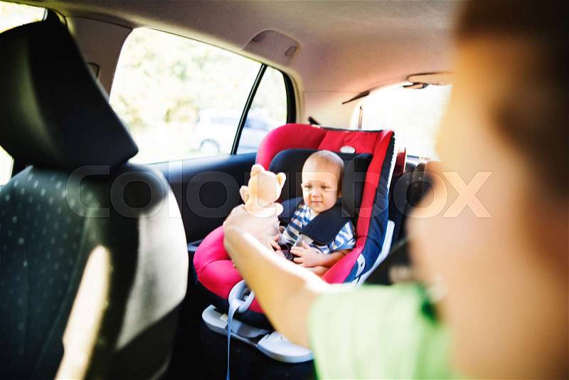 Young mother with her little son in the car. Baby boy sitting in a car seat, unrecognizable woman giving him a toy, stock photo