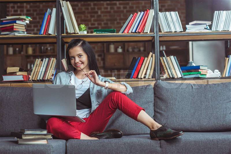 Girl studying with laptop and books while sitting on sofa and smiling at camera, stock photo