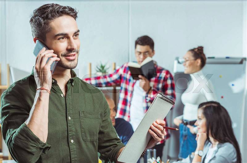 Smiling young handsome man with clipboard talking on smartphone while friends talking behind, stock photo
