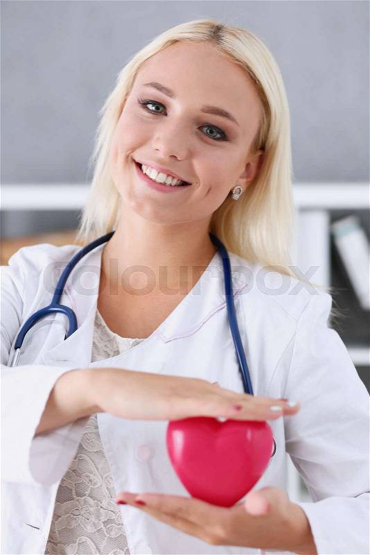 Beautiful smiling blond female doctor hold in arms red toy heart closeup. Cardio therapeutist student education CPR 911 life save physician make cardiac physical pulse rate measure arrhythmia, stock photo