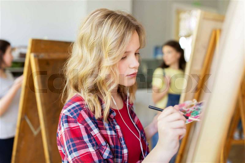 Art school, creativity and people concept - student girl or artist with earphones, easel and paint brush painting at studio, stock photo