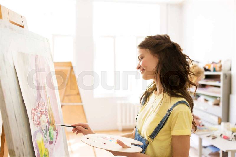 Art school, creativity and people concept - student girl or young woman artist with easel, palette and paint brush painting still life picture at studio, stock photo