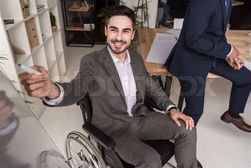 Smiling disabled businessman in wheelchair pointing at whiteboard during business meeting with colleagues, stock photo