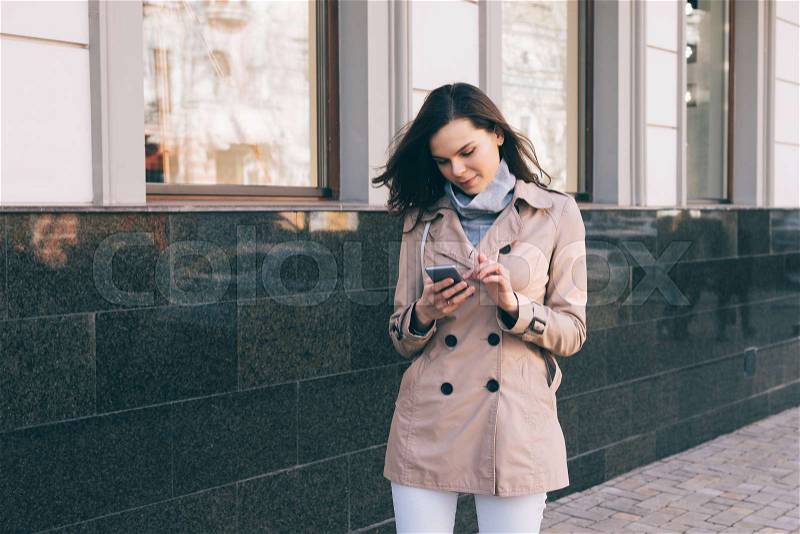 Young slim woman in a beige coat walks down the street and checks the mobile phone, stock photo