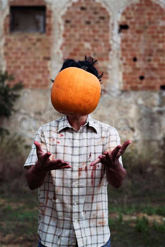 Closeup of a scary man wearing ragged and dirty clothes with blood stains throwing a pumpkin up in the air in front of an abandoned house, stock photo