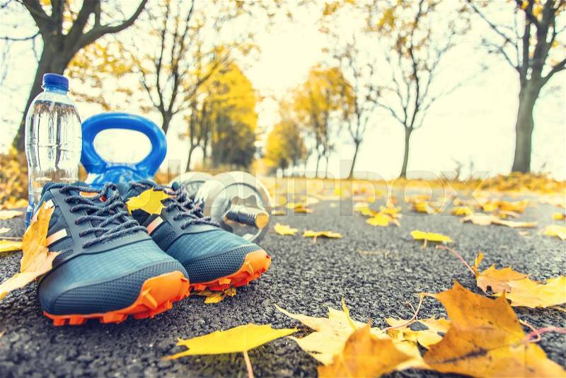 Pair of blue sport shoes water and dumbbells laid on a path in a tree autumn alley with maple leaves - accessories for run exercise or workout activity, stock photo