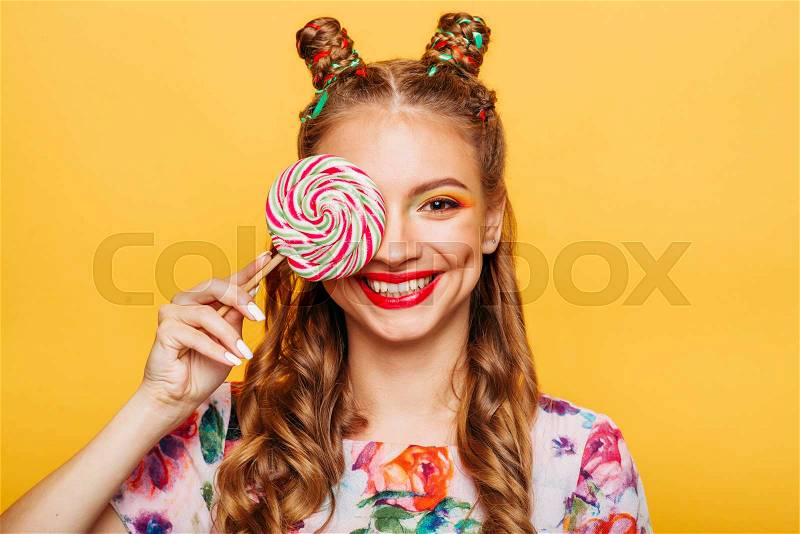 Beautiful young woman holds in hand candy, emotion of indignation on her face. Stylish girl in summer colorful dress. Studio portrait, yellow wall on background, stock photo
