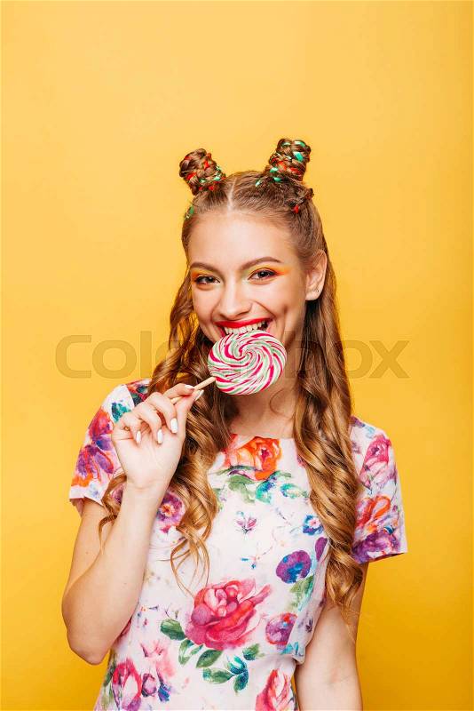 Beautiful young woman with playful look eating candy and smiling. Stylish girl with blonde curly hair. Portrait of attractive lady with big lollypop, yellow wall on background, stock photo