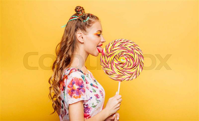 Beautiful young woman with playful look eating huge candy and smiling. Stylish girl with blonde curly hair. Portrait of attractive lady with big lollypop, yellow wall on background, stock photo