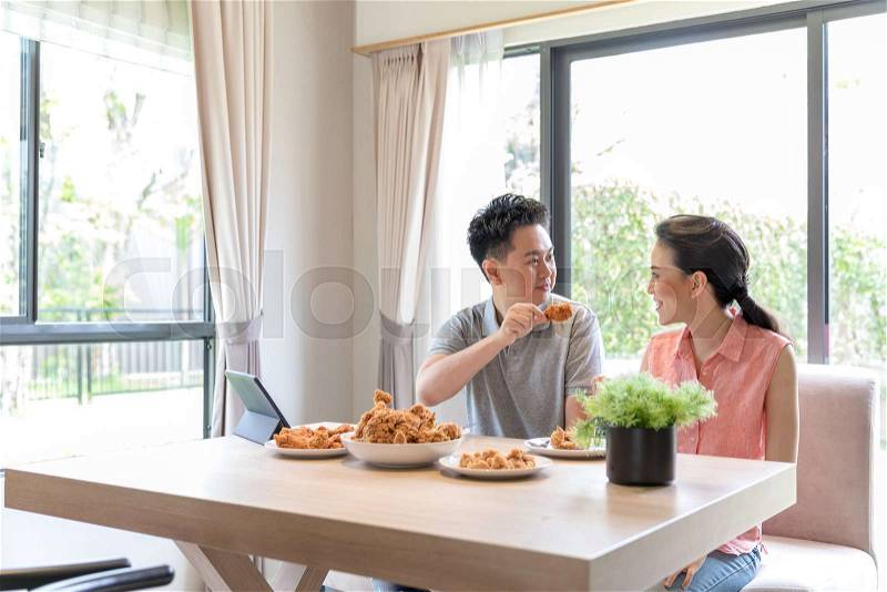 Young Asian Couples eating fried chicken together in living room of contemporary house for modern lifestyle concept, stock photo