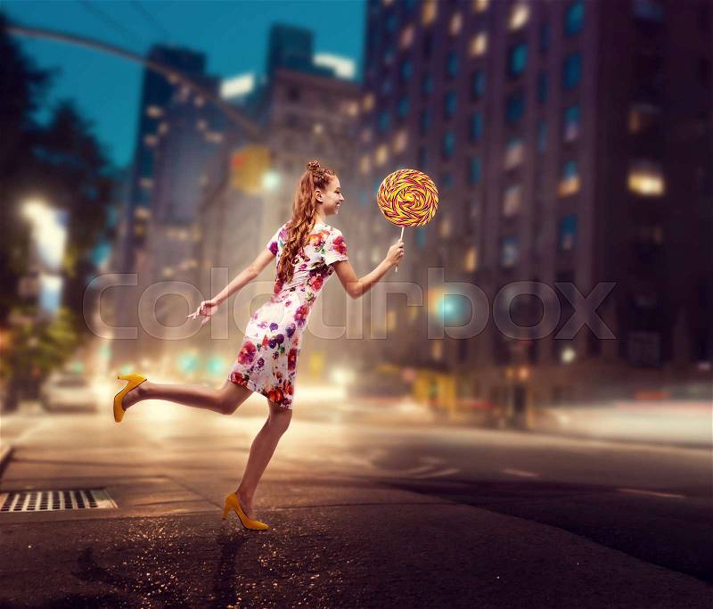 Beautiful young woman run with huge lollipop in her hand. Stylish girl with blonde curly hair. Stylish girl in colorful summer dress, night city on background, stock photo