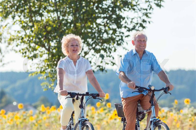 Active elderly couple wearing summer casual clothes while riding bicycles together in the countryside, stock photo