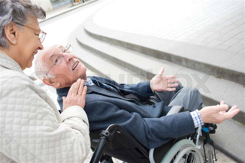 Senior woman pushing her disabled husband in the street, stock photo
