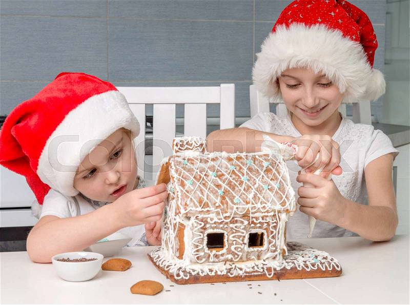 Two cute little sisters in santa hat making biscuit house with cream, stock photo