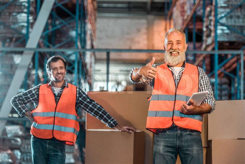 Cheerful warehouse workers with digital tablet smiling at camera and showing thumb up, stock photo