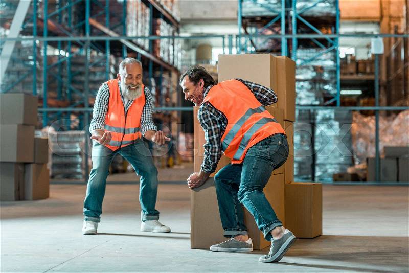 Smiling bearded worker looking at colleague moving boxes in warehouse, stock photo