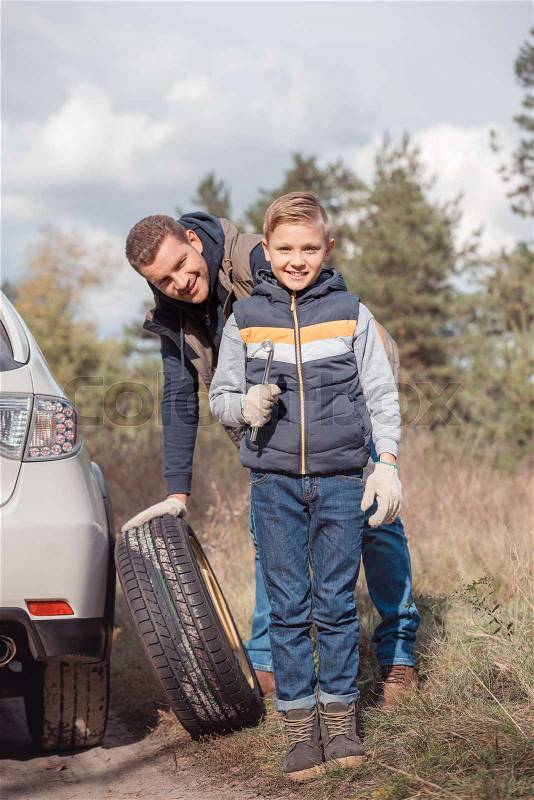 Father and son smiling at camera while changing car wheel together, stock photo