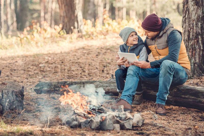 Happy father and son using digital tablet while sitting on log in autumn forest, stock photo