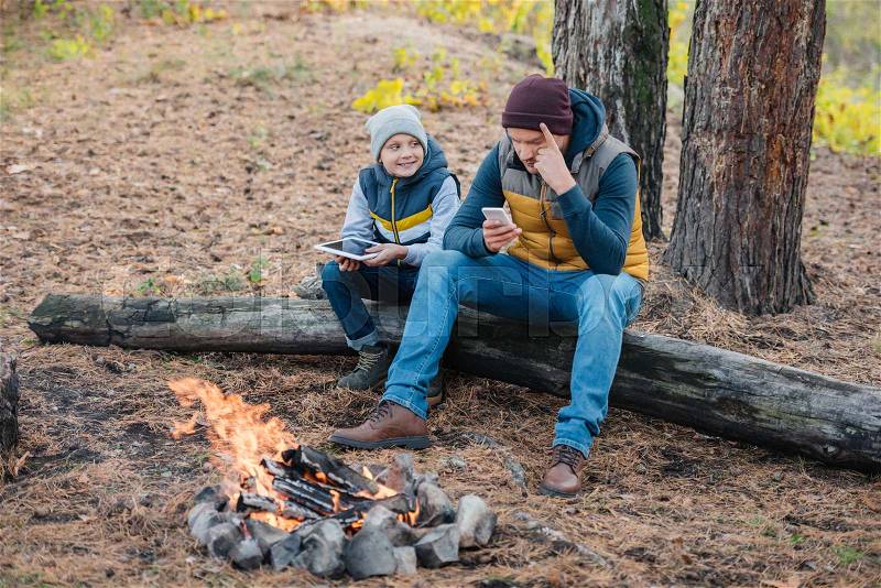 Father and son using digital devices while sitting on log in forest, stock photo