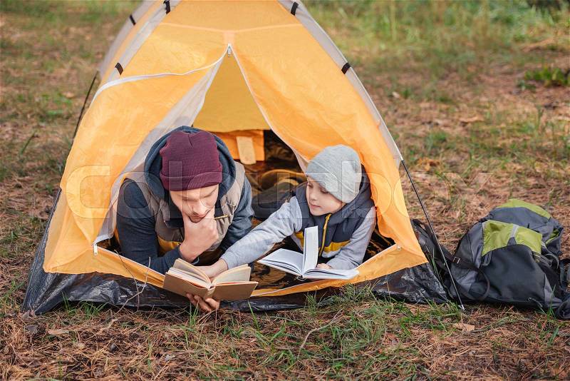 Father and son reading books while lying in tent in autumn forest, stock photo