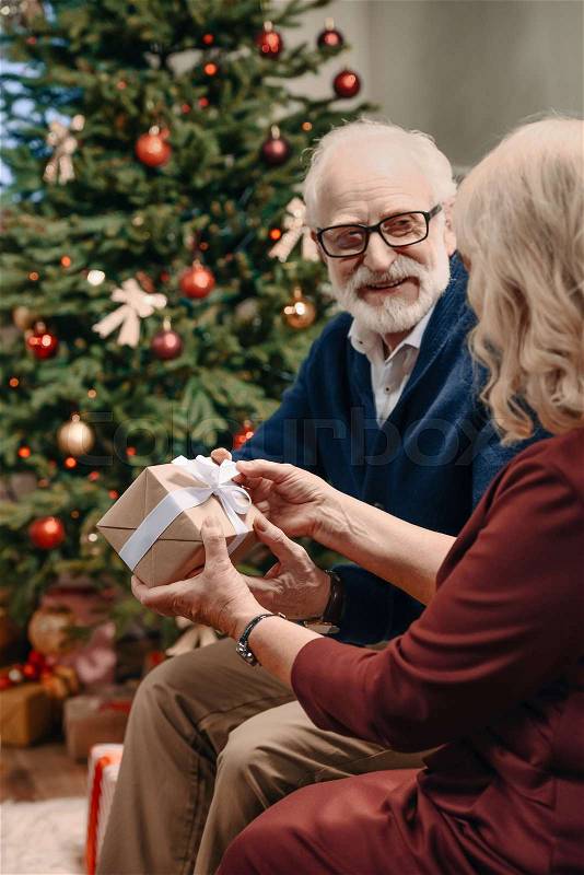 Happy senior man presenting christmas gift to wife in front of beautiful christmas tree, stock photo