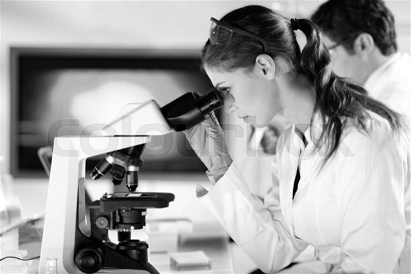Life scientists researching in laboratory. Attractive female young scientist and her post doctoral supervisor microscoping in their working environment. Healthcare and biotechnology. Black and white, stock photo