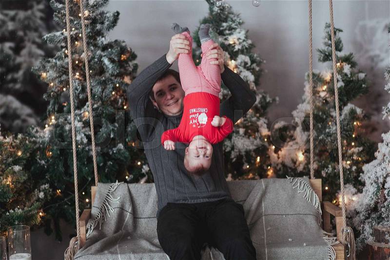Father and 1 year old son fooling around in front of Christmas tree sitting on wooden swing in studio. Dad hold child turned upside down, stock photo