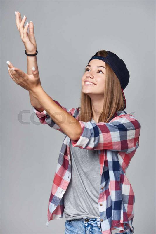 Smiling teen girl with raised hands to catch something falling from the above over gray background, stock photo