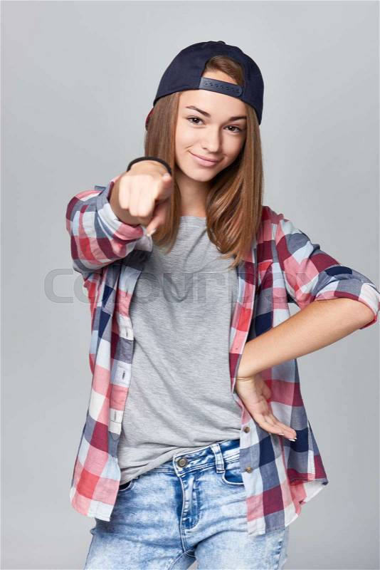 Smiling teen girl pointing at camera with cool attitude, over grey background, stock photo