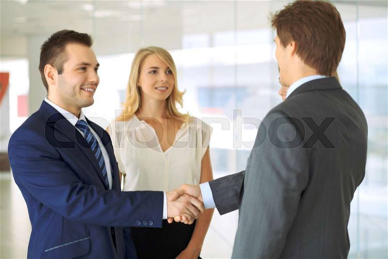 Businessmen shaking hands. Two confident businessmen shaking hands and smiling while standing at office together with people in the background, stock photo