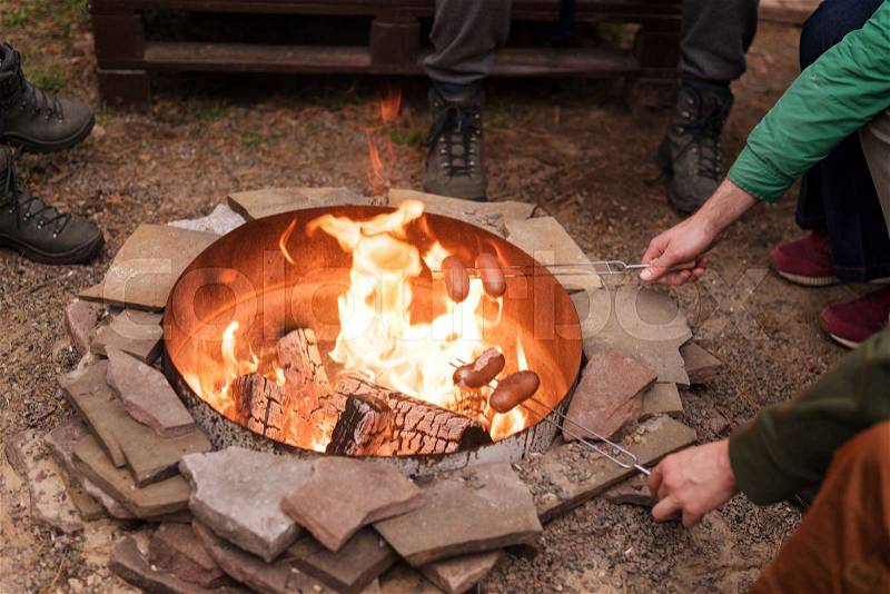 Grilling sausages over a campfire, campers roasting sausages on toasting forks. Fire place, friends, tourists are sitting near the flame, stock photo