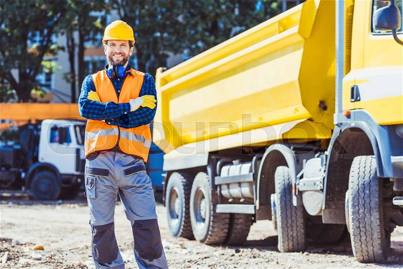 Smiling worker in reflective vest and hardhat posing with arms crossed in front of yellow tip truck at construction site, stock photo