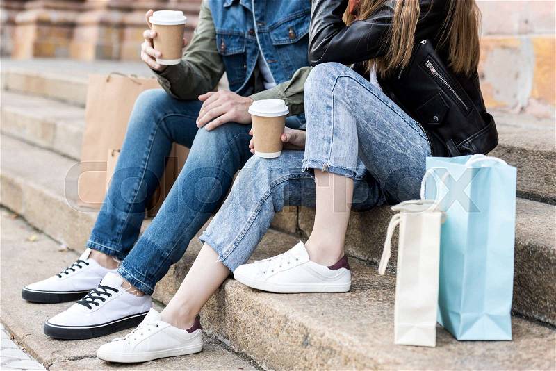 Partial view of couple with shopping bags and coffee to go sitting on stairs on street , stock photo