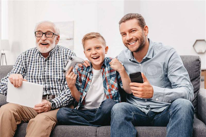 Father, son and grandfather sitting together on couch in living room holding digital tablet, smartphone and gamepad, stock photo