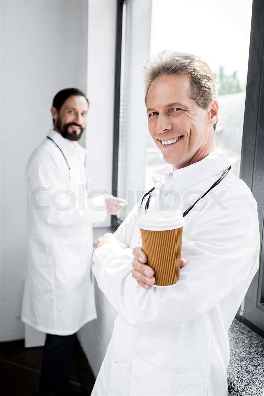 Handsome male doctor holding disposable coffee cup and smiling at camera, stock photo
