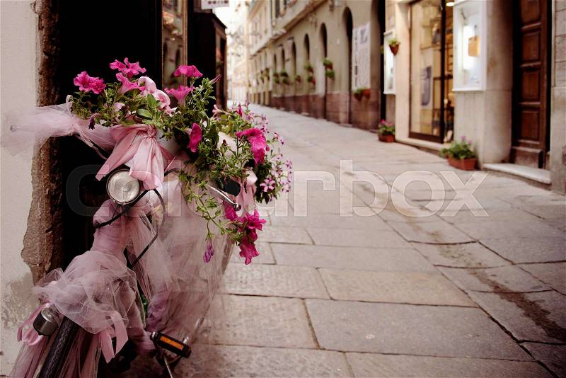 A pedestrian street in the old town of Alghero, Sardinia, Italy, with a bicycle ornamented with flowers and pink fabrics and ribbons parked in a wall, stock photo