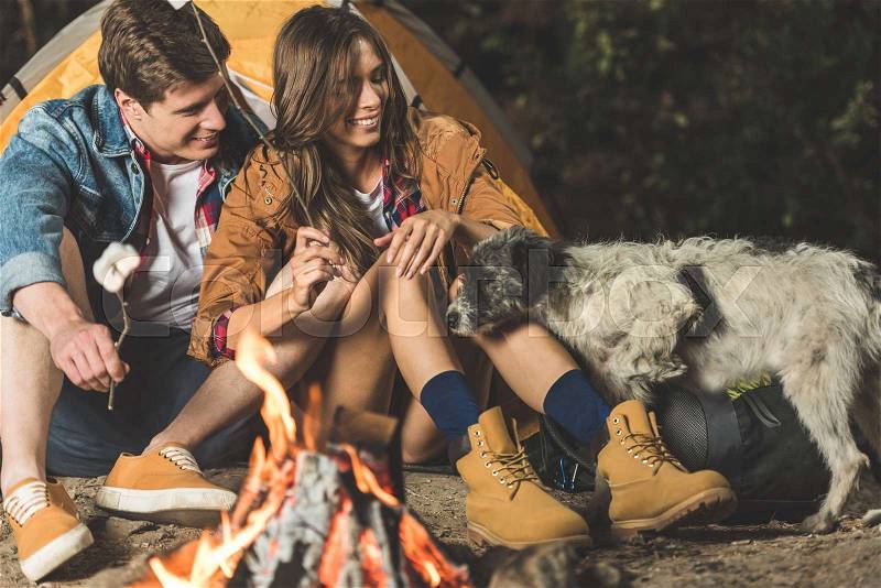 Young happy couple on hiking trip with old dog roasting marshmallow , stock photo