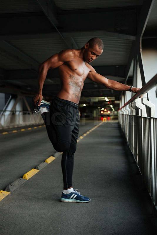 Full length portrait of a motivated half naked african sportsman doing stretching exercises while leaning on a handrail indoors, stock photo