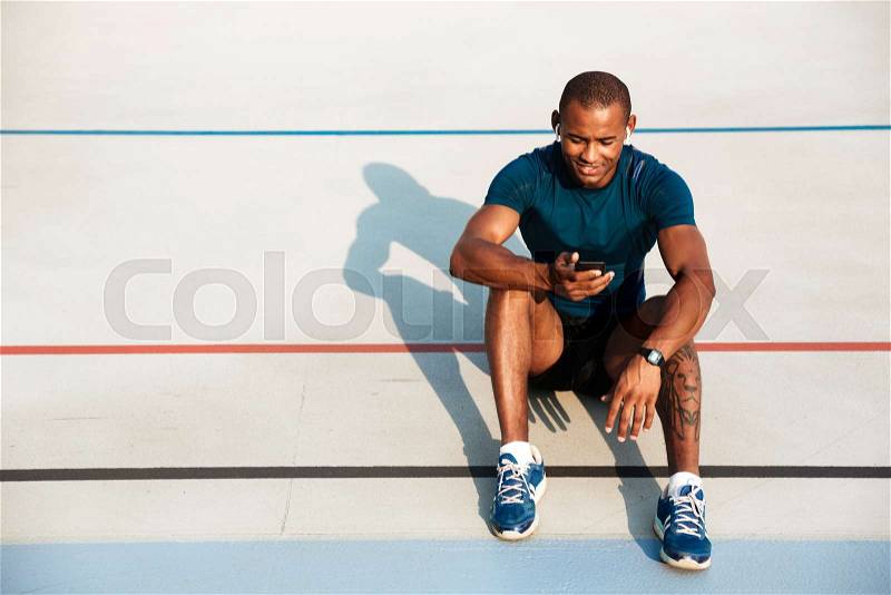 Portrait of a smiling young african fitness man in earphones resting while sitting on a ground at the track field outdoors, stock photo