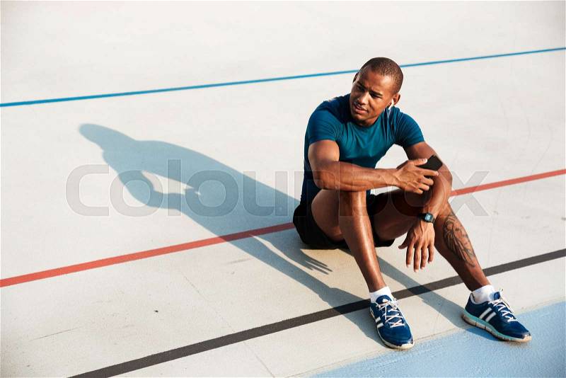 Portrait of a young motivated african fitness man in earphones resting while sitting on a ground at the track field outdoors and looking away, stock photo