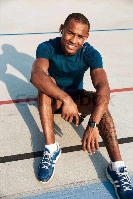 Portrait of a smiling motivated african fitness man in earphones resting while sitting on a ground at the track field outdoors and looking at camera, stock photo