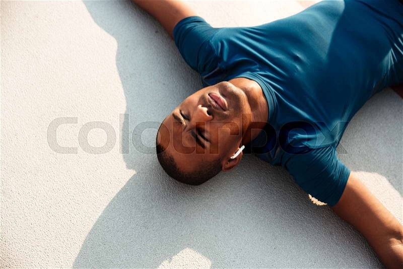 Close up portrait of a exhausted young african fitness man in earphones resting while lying on a ground at the track field outdoors, stock photo