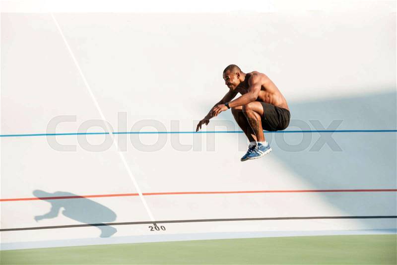 Full length portrait of a half naked fit african sportsman doing a high jump at the track field outdoors, stock photo