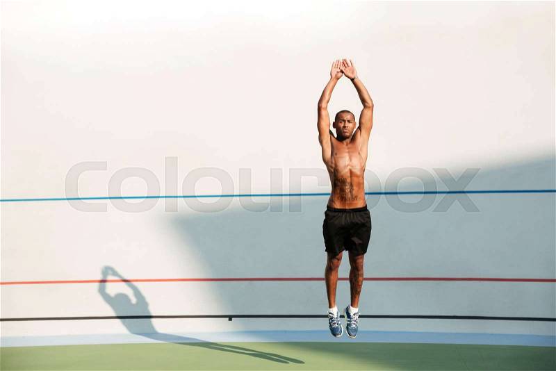 Full length portrait of a half naked african fitness man jumping at the track field outdoors, stock photo